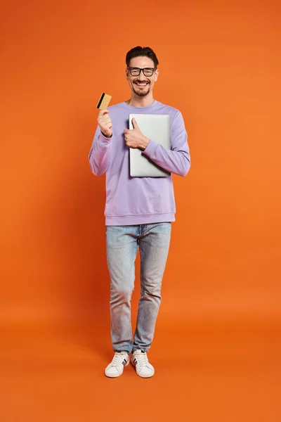 stock image happy man in glasses standing with credit card and laptop on orange background, online shopping