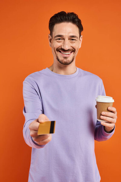 happy man in purple sweater holding coffee to go and offering credit card on orange background