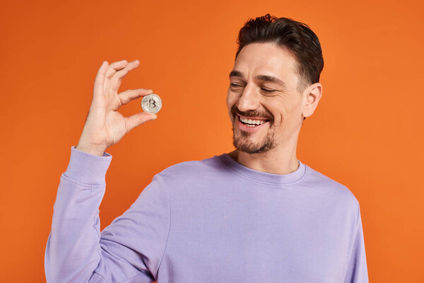 счастливый бородатый мужчина держит биткойн и улыбается на оранжевом фоне, криптовалютные инвестиции