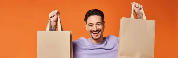 stock image happy bearded man in casual attire holding shopping bags on orange background, consumerism banner