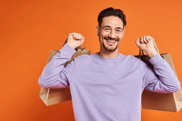 stock image positive bearded man in casual attire holding shopping bags on orange background, consumerism