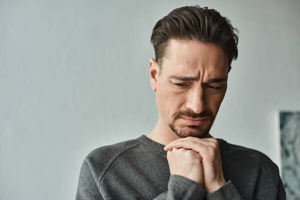 грустный и бородатый мужчина в повседневной домашней одежде стоя с руками возле лица на сером фоне, депрессия
