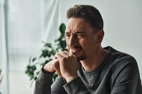 расстроенный бородатый мужчина в повседневной одежде дома чувствует себя нездоровым и хмурясь, глядя в сторону дома