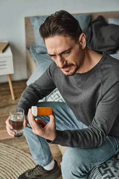 stock image bearded man holding glass of water while looking at medication in bottle and sitting on bed