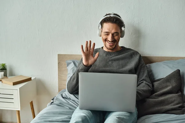 cheerful man in wireless headphones using laptop while waving hand during video call, freelance