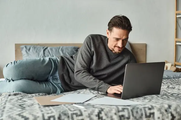 stock image focused and bearded man using laptop while working remotely from home in bedroom, documents