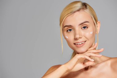 Sarışın ve yanaklarında krem ve krem olan çekici bir kız. Gri arkaplanlı bir elle çeneye dokunuyor.