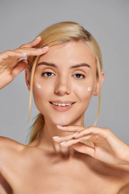 portrait alluring girl in her 20s with her hands and cream on cheeks against grey background clipart