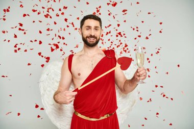 bearded man in cupid costume with heart-shaped arrow and champagne under red confetti on grey clipart