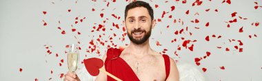 cheerful bearded man dressed as cupid with arrow and champagne under red confetti on grey, banner clipart