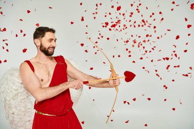 Kırmızı konfeti altında kalp şeklinde okla okçuluk yapan sırıtan sakallı adam Sevgililer Günü