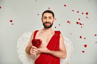Aşk tanrısı gibi giyinmiş mutlu sakallı bir adam. Gri kostümlü, kırmızı kulak şeklinde sevgililer günü hediyeleri var.