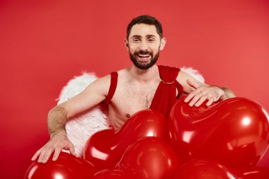 bearded man in cupid costume and wings laughing near heart-shaped balloons on red, st valentines day clipart