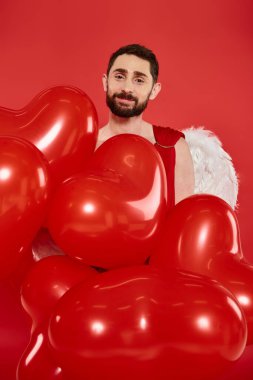 Kalp şeklinde balonları olan sakallı sakallı adam kırmızı sevgililer günü partisinde kameraya bakıyor.