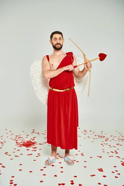 stock image joyful bearded man dressed as cupid archering on grey backdrop, Saint Valentines day costume party