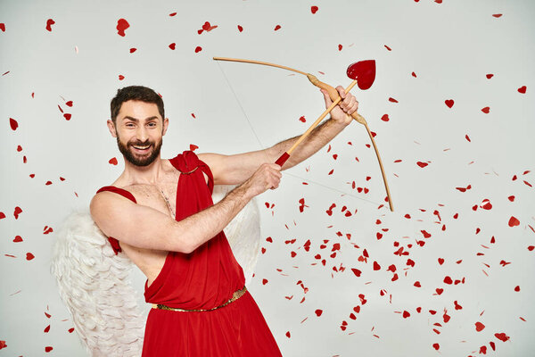 excited bearded cupid man archering under heart-shaped confetti on grey, st valentines party
