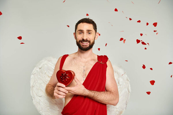 happy bearded man dressed as cupid with red hear-shaped st valentines present on grey, costume party