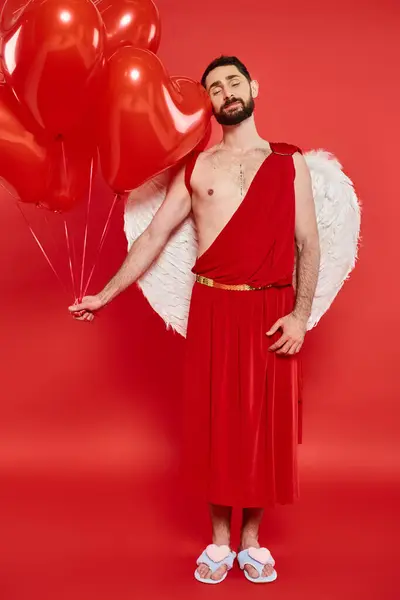 stock image romantic bearded man in cupid costume with closed eyes and red balloons on red, st valentines day