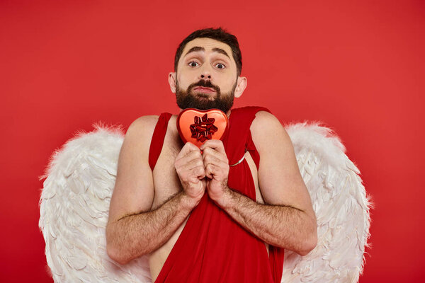 funny man in cupid costume and wings holding heart-shaped gift box and looking at camera on red