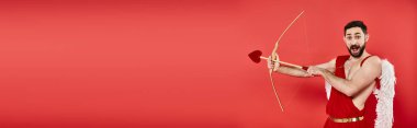 thrilled cupid man with open mouth archering while looking at camera on red, horizontal banner clipart