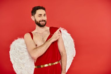 cheerful bearded cupid man looking at camera and pointing away with finger on red, st valentines day clipart