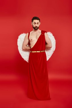 full length of thoughtful bearded man in cupid costume standing with hands behind back on red clipart