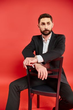 Siyah takım elbiseli karizmatik sakallı iş adamı. Sandalyeye oturuyor ve kırmızı arka plana bakıyor.