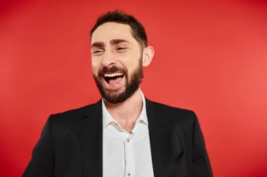 portrait of excited bearded businessman in black suit looking away and laughing on red backdrop clipart