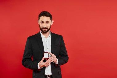 Siyah takım elbiseli gülümseyen sakallı adam kırmızı takı kutusunda yüzüğü gösteriyor.