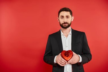 Keskin giyimli sakallı bir adam Sevgililer Günü hediyesini kırmızı üzerinde kalp şeklinde bir hediye kutusunda taşıyor.