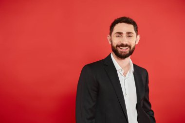 Kırmızı fonda kameraya gülümseyen siyah şık takım elbiseli başarılı sakallı iş adamının portresi.