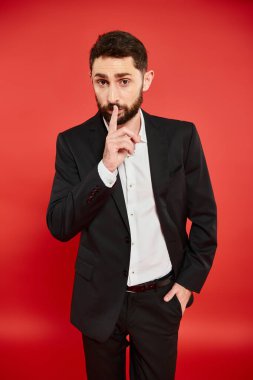 bearded man in black suit showing hush sign while standing with hand in pocket on red backdrop clipart