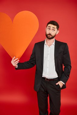 bearded elegant man in black suit with orange paper heart looking away on red, st valentines day clipart