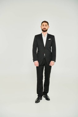 front view of bearded businessman in black elegant suit standing on grey backdrop, full length clipart