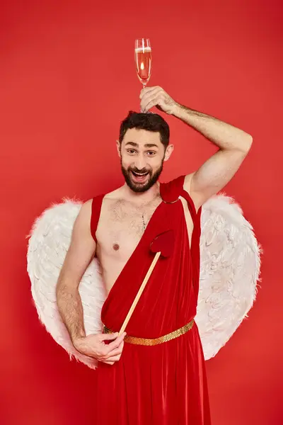 stock image funny and joyful man in cupid costume posing with heart-shaped arrow and champagne glass on head