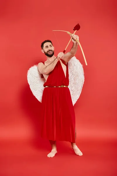 stock image full length of excited man in cupid costume archering on red, Saint Valentines day concept