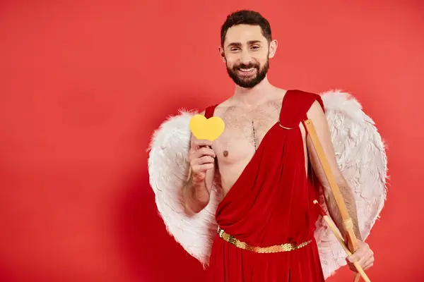 stock image cheerful bearded man in cupid costume with bow and yellow paper heart on red, Saint Valentines day