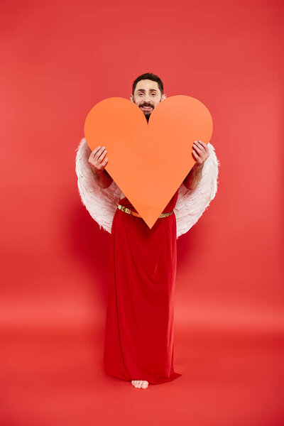 smiling bearded man dressed as cupid holding huge orange heart on red, valentines costume party