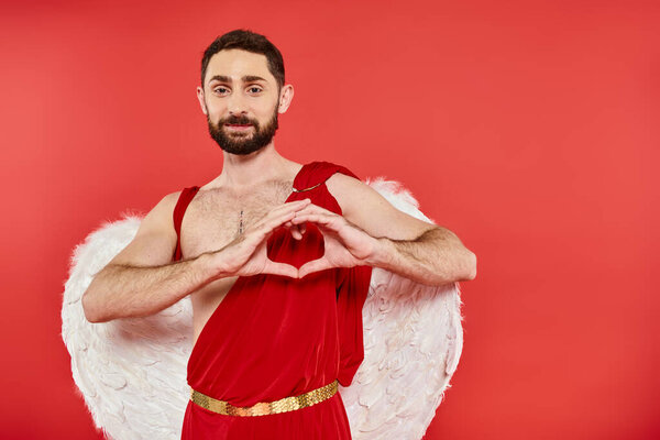 bearded man in costume and wings of cupid showing heart sign with hands and smiling at camera on red