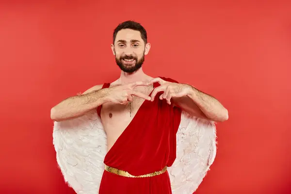 stock image pleased bearded cupid man showing heart sign with fingers and smiling at camera on red backdrop