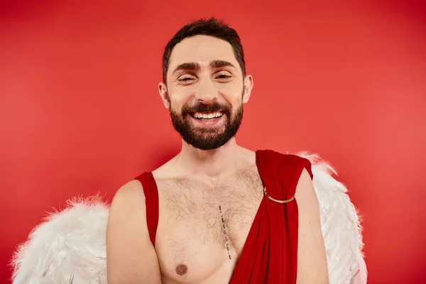 stock image portrait of cheerful bearded man dressed as cupid smiling at camera on red, st valentines party