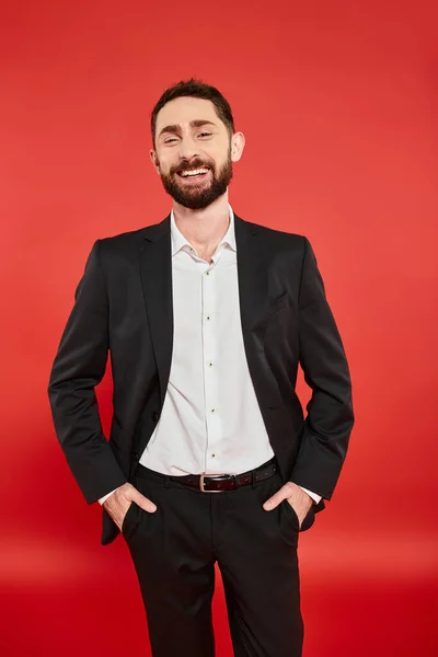 stock image joyful businessman in black elegant suit standing with hands in pockets and smiling at camera on red