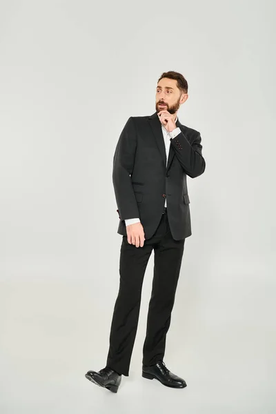 stock image full length of serious elegant businessman in black suit touching beard and looking away on grey