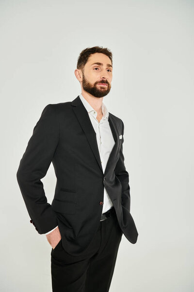confident bearded businessman in black suit holding hands in pockets and looking away on grey