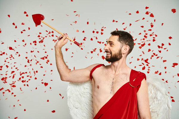cheerful bearded cupid man with heart-shaped arrow under red confetti on grey, st valentines day
