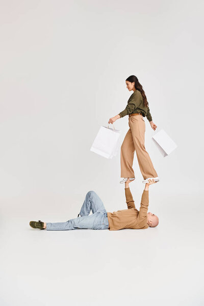 woman in casual wear holding shopping bags and balancing on hands of man, couple of acrobats