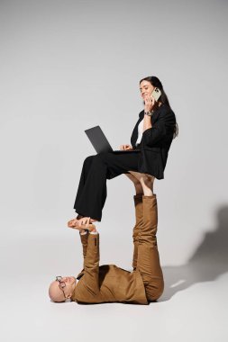 İş kıyafetleri içinde mutlu bir kadın, laptopuyla telefonla konuşuyor ve gri üzerinde bir adamın ayakları üzerinde dengede duruyor.