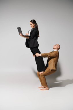 Woman in business attire with laptop balancing on laps of man in suit on grey, couple of acrobats clipart