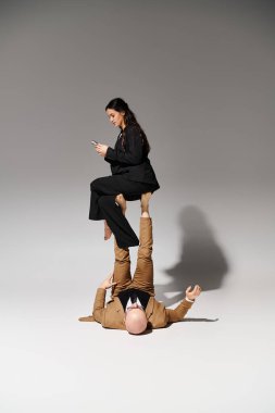 young businesswoman with smartphone balancing on feet of man in suit on grey, couple of acrobats clipart