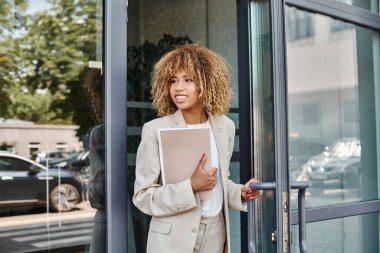 Kıvırcık saçlı, klasörlü ve ofis binasından çıkan mutlu Afro-Amerikan iş kadını.